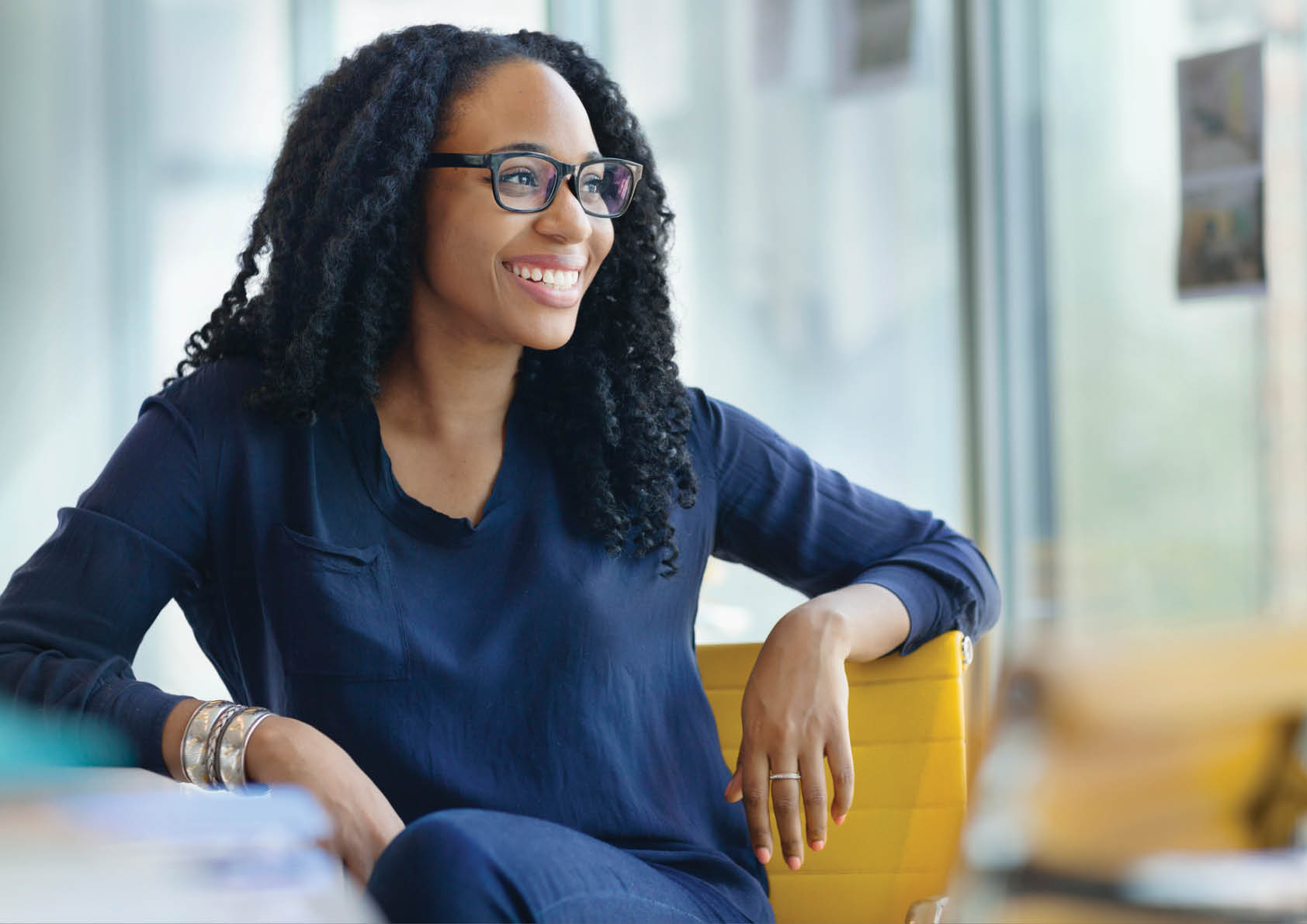 Portrait of young Black female woman designer owner entrepreneur small business new hire intern smiling proud dedicated passionate looking at camera sitting at desk in creative design studio office startup company contemporary modern daylight bright sunny natural light curly hair eyeglasses