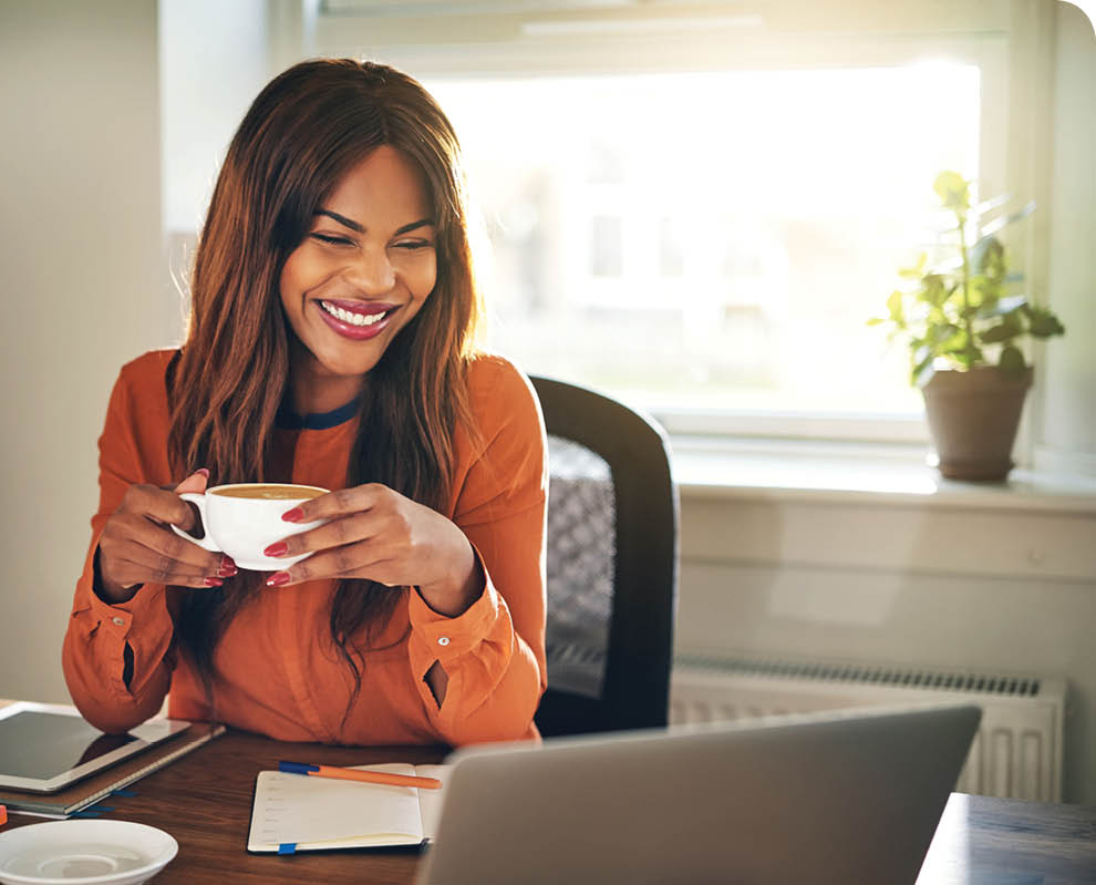 Laughing young African female entrepreneur drinking a cup of coffee and working on a laptop while sitting at a desk in her home office
