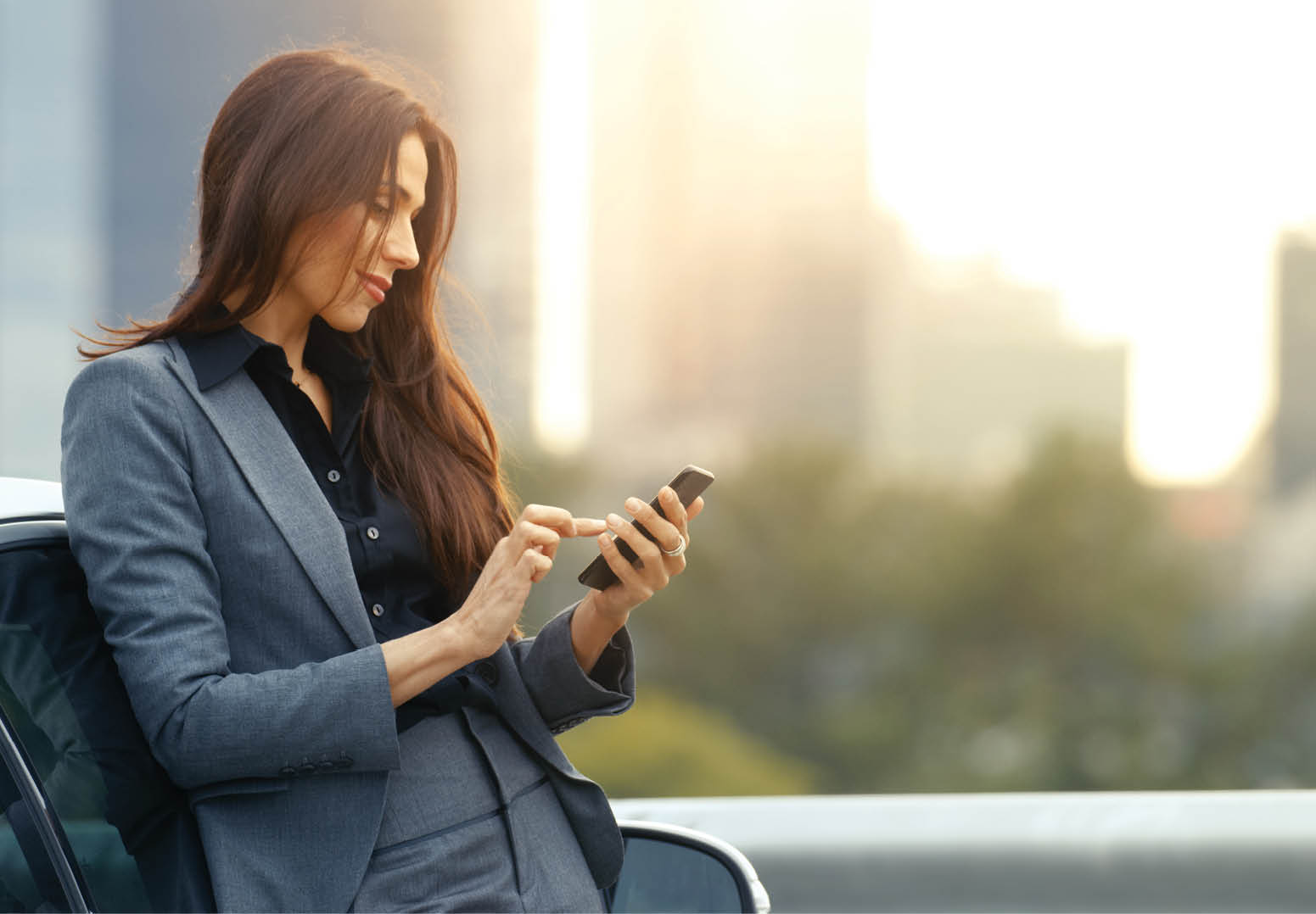 Business Woman Uses Smartphone While Leaning on Her Premium Class Car  Big City with Skyscrapers in the Background 
