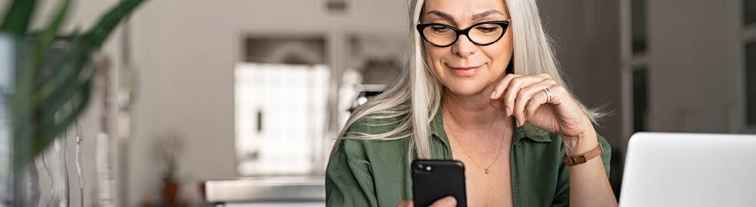 Happy senior woman using mobile phone while working at home with laptop  Smiling cool old woman with white hair wearing eyeglasses sitting on chair at table and messaging with smartphone  Beautiful stylish elderly lady browsing site on cellphone 